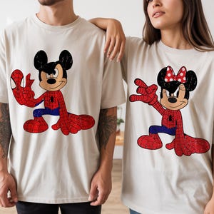 May include: Two beige short-sleeve t-shirts featuring graphic prints. The left shirt displays Mickey Mouse in a red and blue spider-themed suit. The right shirt displays Minnie Mouse in a matching red and blue spider-themed suit with a red bow.