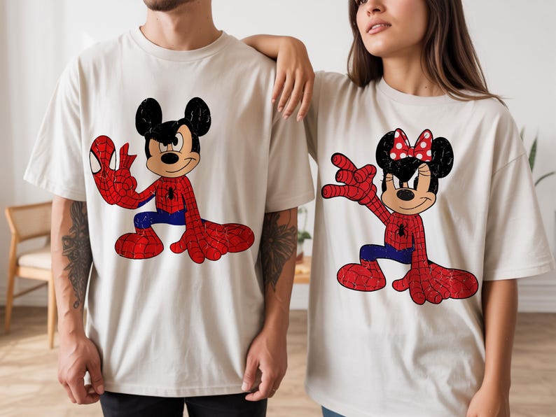 May include: Two beige short-sleeve t-shirts featuring graphic prints. The left shirt displays Mickey Mouse in a red and blue spider-themed suit. The right shirt displays Minnie Mouse in a matching red and blue spider-themed suit with a red bow.