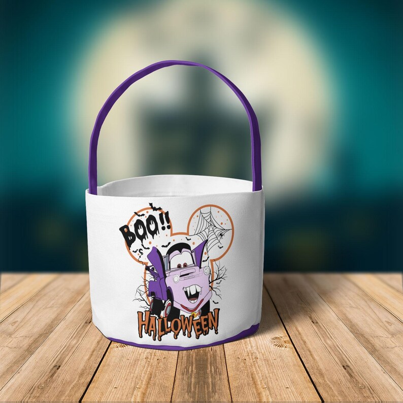 May include: A white Halloween bucket with a purple handle and base. The front features a graphic of a purple vampire car inside a mouse-ear silhouette, with a spider web, bats, and the text BOO!! and HALLOWEEN printed in black and orange.