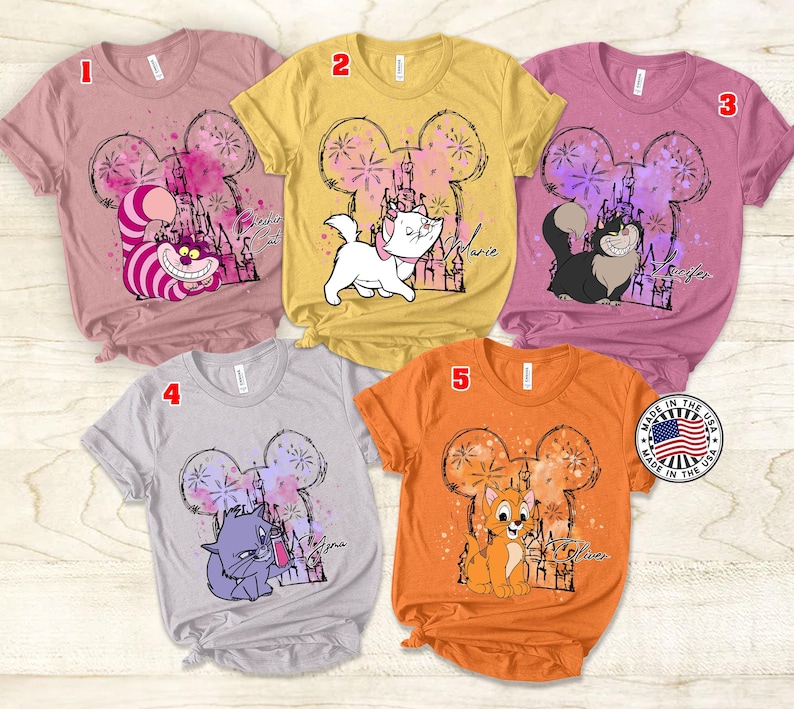 Puede incluir: Cinco camisetas diferentes con una cabeza de rat&oacute;n de dibujos animados con un castillo y fuegos artificiales en el fondo. Cada camiseta presenta un personaje diferente de Disney: Marie, Figaro, Duquesa, Berlioz y Toulouse. Las camisetas son de varios colores, incluyendo rosa, amarillo, gris y naranja.