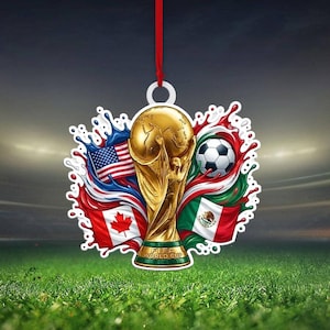 May include: A decorative hanging ornament featuring the gold FIFA World Cup trophy at the center, surrounded by the flags of the United States, Canada, and Mexico, a soccer ball, and colorful paint splashes, suspended by a thin red ribbon.