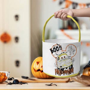 May include: A white cylindrical Halloween bucket with a yellow handle. The bucket features a graphic of a yellow car wrapped in bandages like a mummy, a spider web, and the text "BOO!! HALLOWEEN" printed in black and orange lettering.