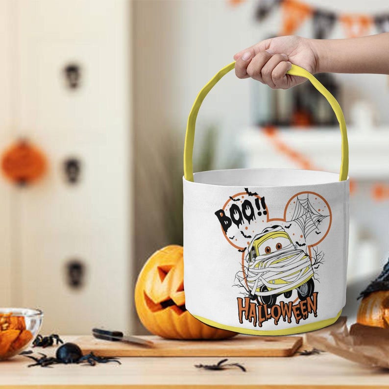 May include: A white cylindrical Halloween bucket with a yellow handle. The bucket features a graphic of a yellow car wrapped in bandages like a mummy, a spider web, and the text "BOO!! HALLOWEEN" printed in black and orange lettering.