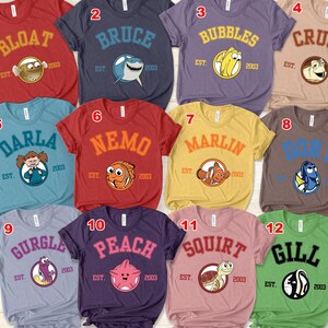 Op de afbeelding: Diverse gekleurde t-shirts met personages uit een populaire animatiefilm. Elk shirt toont de naam van een personage in een universiteitslettertype, een afbeelding van het personage en de tekst "EST. 2003".