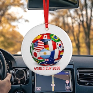 May include: A round white ceramic ornament hanging from a red ribbon, featuring a soccer ball graphic composed of various national flags. The text WORLD CUP 2026 is printed in red capital letters at the bottom of the ornament surface.