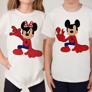 May include: Two white short-sleeved t-shirts featuring graphic prints. The left shirt displays a character with a red polka-dot bow wearing a red and blue spider-web patterned suit. The right shirt shows a character in a similar spider-web suit.