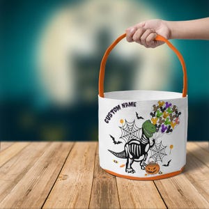 Op de afbeelding: Witte Halloween-emmer met een oranje handvat en rand. De emmer heeft een dinosaurus skelet afbeelding, spinnenwebben, vleermuizen en een pompoen. De tekst op de emmer luidt "CUSTOM NAME".