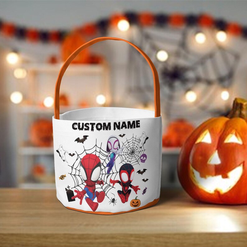 May include: A white cylindrical Halloween candy bucket with an orange handle. The exterior features a graphic of three cartoon superheroes, spiderwebs, bats, ghosts, and a pumpkin. The text CUSTOM NAME is printed in black above the characters.