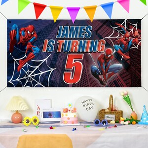 Peut inclure: Une banni&egrave;re d'anniversaire avec des graphismes de Spider-Man et le texte "JAMES IS TURNING 5" sur fond de paysage urbain. Des fanions color&eacute;s sont suspendus au-dessus. Une table est dress&eacute;e avec un g&acirc;teau, des ballons et des d&eacute;corations.