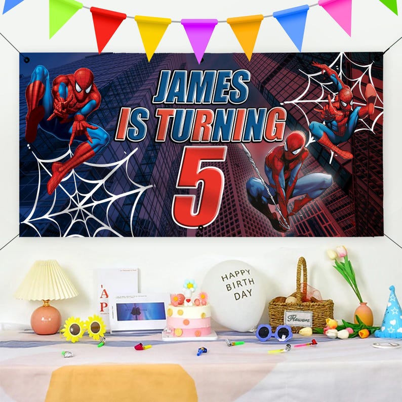 Peut inclure: Une banni&egrave;re d'anniversaire avec des graphismes de Spider-Man et le texte "JAMES IS TURNING 5" sur fond de paysage urbain. Des fanions color&eacute;s sont suspendus au-dessus. Une table est dress&eacute;e avec un g&acirc;teau, des ballons et des d&eacute;corations.