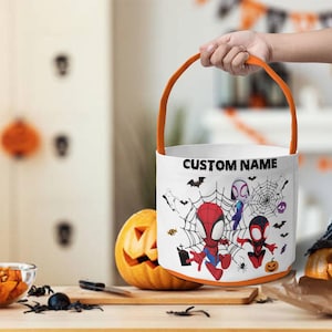 May include: A white cylindrical Halloween candy bucket with an orange handle. The exterior features a graphic print of cartoon superheroes, spiderwebs, bats, and ghosts. The text CUSTOM NAME is printed in black letters at the top of the bucket.