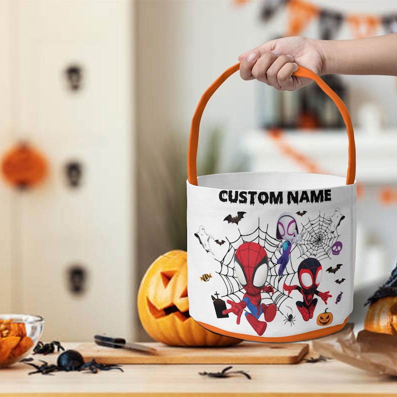 May include: A white cylindrical Halloween candy bucket with an orange handle. The exterior features a graphic print of cartoon superheroes, spiderwebs, bats, and ghosts. The text CUSTOM NAME is printed in black letters at the top of the bucket.