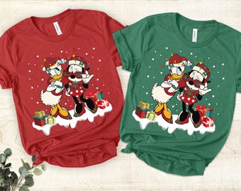 May include: Red and green t-shirts featuring a festive cartoon design of Donald Duck and Minnie Mouse. They are wearing Christmas hats and sunglasses, standing on a snowy platform with wrapped gifts. The shirts are short-sleeved.