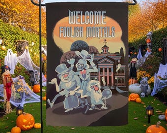Op de afbeelding: Een rechthoekige tuin vlag met een zwarte achtergrond en de tekst "WELCOME FOOLISH MORTALS" in het wit. De illustratie toont drie cartoon spoken voor een landhuis, met een grote oranje maan achter de tekst en het gebouw.