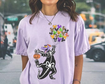 May include: A light purple short-sleeve t-shirt featuring a graphic of a purple dragon wearing a white skeleton costume. The dragon holds a bunch of colourful balloons shaped like mouse heads and a small orange jack-o'-lantern candy bucket.