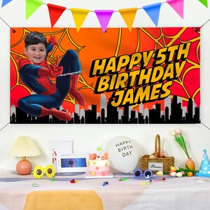 以下が含まれることがあります： 「Happy 5th Birthday James」と書かれた赤と青のスパイダーマンの誕生日バナー。バナーには、街並みを背景にスイングするスパイダーマンの漫画の画像が描かれています。