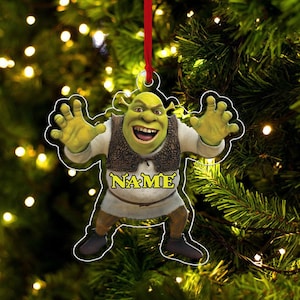 Pode incluir: Um enfeite de Natal com o personagem Shrek com os braços estendidos. O enfeite é verde, castanho e branco, com a palavra "NAME" em letras douradas. Pendura-se de uma fita vermelha contra uma árvore de Natal.
