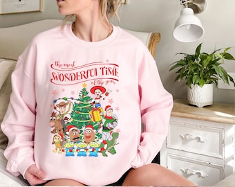 May include: A light pink crewneck sweatshirt featuring a festive graphic of Toy Story characters around a Christmas tree. The text reads, "the most WONDERFUL TIME of the year," with illustrations of Woody, Buzz Lightyear, Jessie, and others wearing Santa hats amidst red snowflakes.