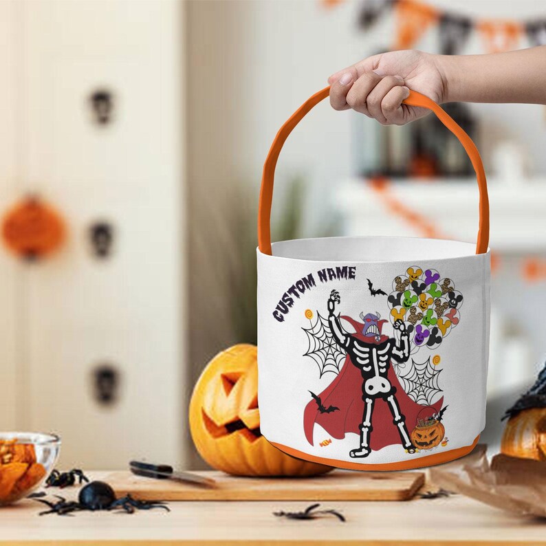 Op de afbeelding: Witte Halloween-emmer met een oranje handvat en rand. De emmer heeft een skeletpersonage met een rode cape, die een cluster van kleurrijke stripfiguren vasthoudt. De woorden "CUSTOM NAME" staan op de zijkant. Een uitgesneden pompoen staat in de buurt.