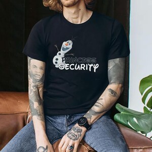 以下が含まれることがあります： 黒いTシャツ。漫画のキャラクター、オラフと「PRINCESS SECURITY」の文字が白でプリントされています。デザインは、笑顔の雪だるまが特徴です。このTシャツはクルーネックで、柔らかい素材で作られています。