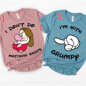 Puede incluir: Dos camisetas a juego, una rosa y otra azul. La camiseta rosa presenta una imagen de dibujos animados de Grumpy de Blancanieves y el texto "I don't do matching shirts". La camiseta azul presenta una mano de dibujos animados apuntando hacia la derecha y el texto "I'm with Grumpy".