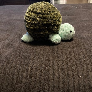 May include: A crocheted green turtle toy with a light green head and legs. The turtle is sitting on a dark gray surface.