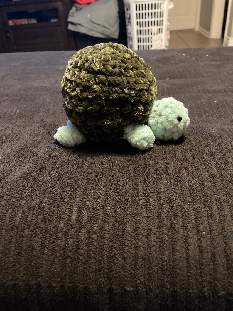 May include: A crocheted green turtle toy with a light green head and legs. The turtle is sitting on a dark gray surface.