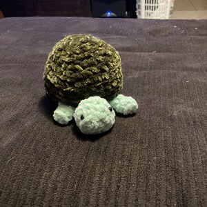 May include: A green and light green crocheted turtle plush toy. The turtle has a round shell and small, round legs.