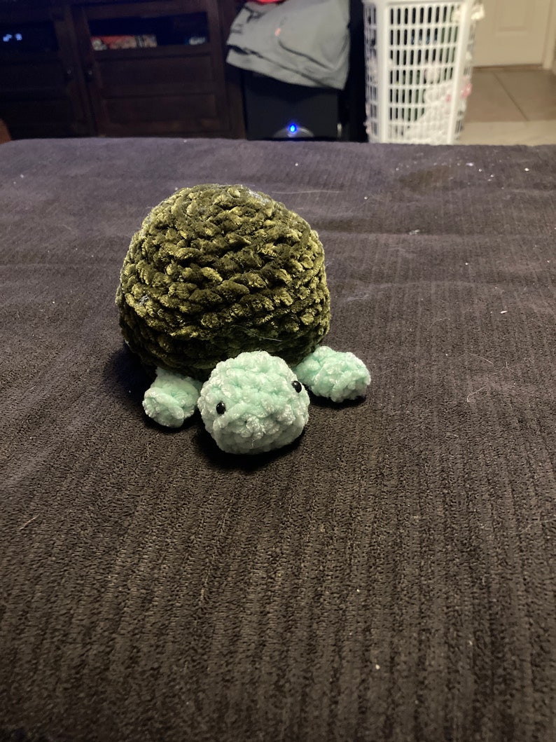 May include: A green and light green crocheted turtle plush toy. The turtle has a round shell and small, round legs.