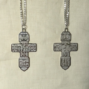 Może przedstawiać: Dwa srebrne łańcuszki z srebrnymi krzyżami. Każdy krzyż ma szczegółowy wzór z postaciami religijnymi i tekstem w obcym języku.