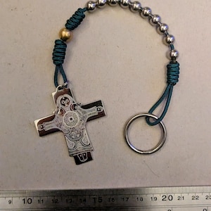 Könnte beinhalten: Ein silberfarbener Kreuzanhänger mit aufwendigen Gravuren, verbunden mit einer Kette aus kleinen, silberfarbenen Perlen. Das Kreuz ist an einem türkisblauen Band und einem silbernen Schlüsselring befestigt. Das Kreuz ist etwa 5 cm lang.