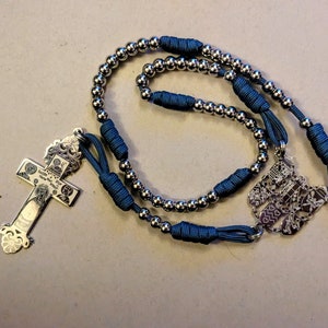 May include: A silver rosary with a silver cross and a silver charm. The rosary is made of small silver beads and a dark blue cord.