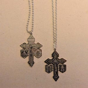 Puede incluir: Dos colgantes de cruz plateados con cadenas de bolas. Cada cruz presenta una intrincada iconografía religiosa, incluyendo representaciones de Jesucristo y santos. Los colgantes están detallados con texto grabado y elementos decorativos.