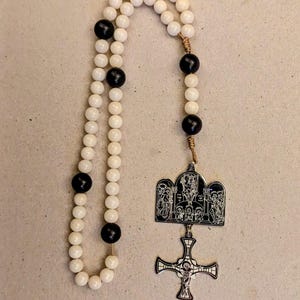Może przedstawiać: Różaniec z koralików w kolorze kości słoniowej z czarnymi koralikami akcentującymi. Różaniec posiada srebrny wisiorek z wizerunkami religijnymi i szczegółowym krucyfiksem. Koraliki są okrągłe i równomiernie rozmieszczone.
