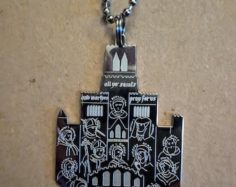 Può includere: Una collana d'argento con un ciondolo a forma di chiesa. Il ciondolo presenta un design dettagliato di una chiesa con molte finestre e un campanile. L'iscrizione sul ciondolo recita "all ye saints and martyrs pray for us".