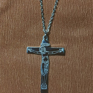 May include: A silver-toned necklace featuring a crucifix pendant. The pendant depicts a detailed image of Jesus Christ on the cross. The chain is a simple link style, and the background is a textured, brown surface.