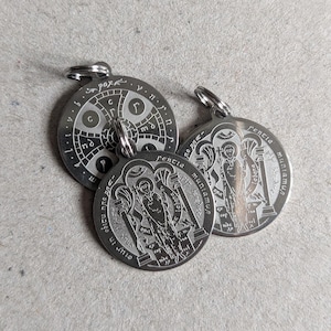 May include: Three circular silver-toned metal pendants featuring engraved religious iconography and Latin text. One pendant displays a cross design with the word pax, while two identical pendants depict a figure standing between pillars with text.