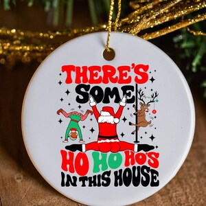 May include: A round, white Christmas ornament with a festive design. The ornament features the text "THERE'S SOME HO HO HO'S IN THIS HOUSE" with illustrations of Santa, an elf, and a reindeer. The ornament is hanging from a gold string.