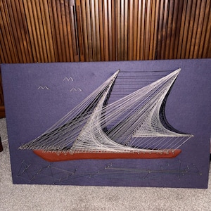 May include: A string art wall piece on a dark purple rectangular board featuring a brown wooden ship hull. White and black threads are woven between nails to form the sails and rigging. Three small metal bird shapes are above the ship.