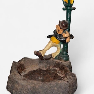 Peut inclure: Un cendrier vintage en fonte représentant une figurine appuyée contre un lampadaire vert. La figurine porte un chapeau marron, une veste noire, une chemise jaune et un pantalon beige. Le cendrier est de forme rectangulaire, sombre et texturé.