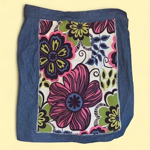 May include: A denim skirt with a large floral pattern. The skirt features a rectangular panel of fabric with large pink, purple, blue, and green flowers on a white background, framed by blue denim.