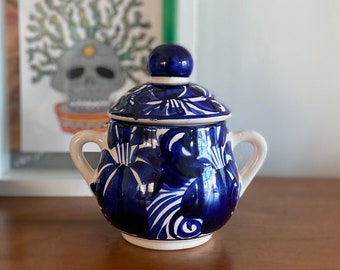May include: A blue and white ceramic sugar bowl with a lid. The bowl has a floral design and is decorated with blue and white paint.