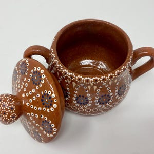 May include: A brown ceramic pot with a lid, both decorated with white and blue dots in a circular pattern. The pot has two handles and a rounded body.