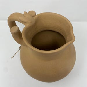 May include: A terracotta pitcher with a unique handle and a slightly flared lip. The pitcher is a light brown color and has a smooth, matte finish.