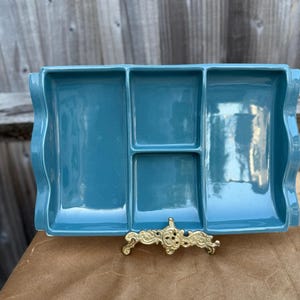 May include: A rectangular teal ceramic serving tray with three distinct compartments and wavy side edges. The tray is supported by a decorative, ornate gold-coloured metal stand featuring intricate scrollwork patterns along its base.