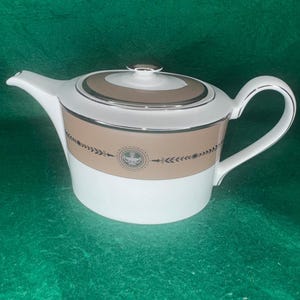 Può includere: Una teiera in porcellana bianca con un'ampia fascia color marrone chiaro attorno al centro, accentuata da un bordo argentato e un medaglione circolare centrale contenente un motivo a urna verde. Il coperchio ha una fascia abbinata e un pomello.