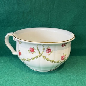 May include: A white ceramic chamber pot featuring a scalloped body and a gold-trimmed rim. The exterior is decorated with a repeating pattern of pink rose clusters connected by green leafy garlands, and it includes a single side handle.