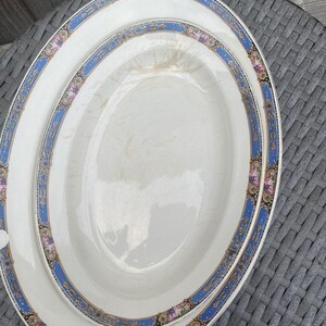 May include: Two oval ceramic serving platters with a cream-colored center and a decorative border featuring a vibrant blue band, gold filigree accents, and small pink floral clusters. The platters have a smooth, glossy finish with visible crazing.