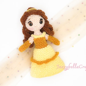May include: A crocheted plush doll of Belle from Beauty and the Beast. The doll is wearing a yellow dress with a brown bodice and has brown hair. The doll is sitting on a white background with a sheer fabric with gold glitter.