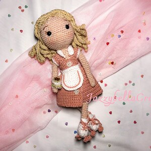 May include: A crocheted doll wearing a pink and white dress with a white apron and roller skates. The doll has blonde hair and brown eyes. The doll is sitting on a pink background with colorful hearts.